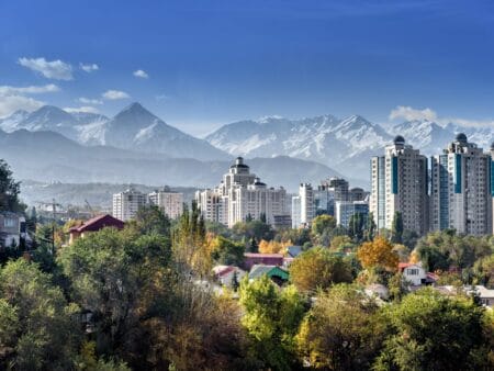 Hochhäuser der Stadt Almaty. Im Hintergrund sind verschneite Berge zu sehen, im Vordergrund farbenfrohe Bäume